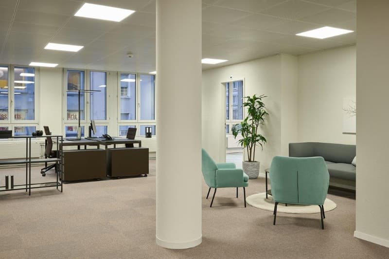 Open office layout with desks, computers, and chairs. Separate seating area with comfortable chairs and a plant. Large windows providing natural light. Bright and modern interior design.
