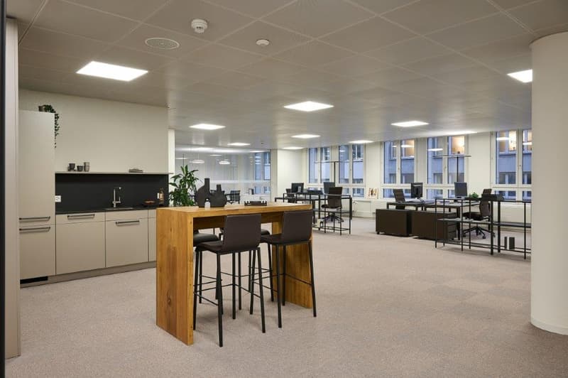 Spacious open-plan office with modern furniture and equipment, including a kitchen area with a dishwasher. The office has large windows, providing natural light, and a mix of individual workstations and collaborative spaces.