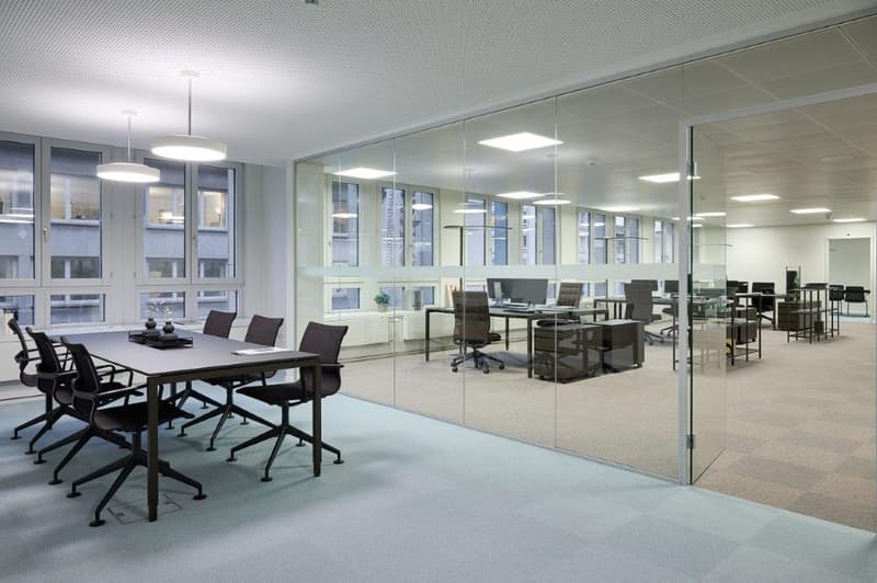 Large open office space with glass walls, conference table, desks, and chairs. The office has a modern and professional design with ample natural lighting.