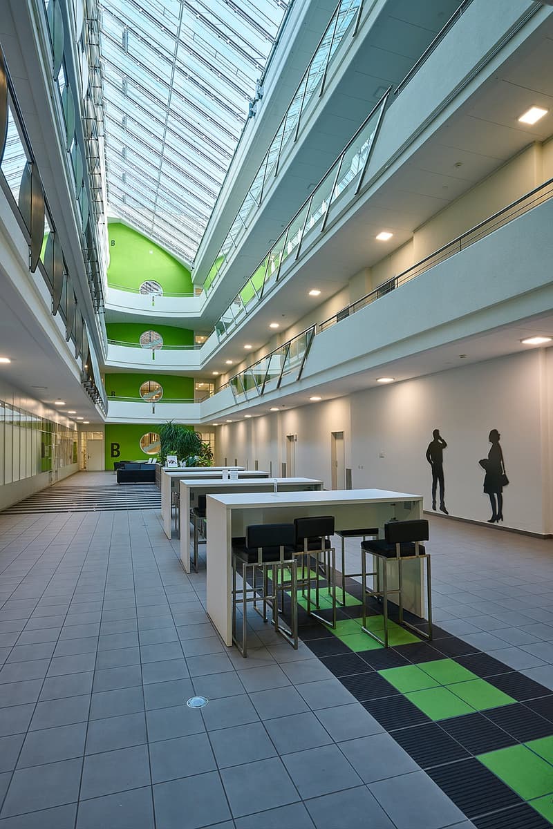The image shows a modern and spacious interior of an office building. The space features a large glass atrium with a slanted roof, allowing natural light to flood the area. The floor is covered with gray and green tiles, creating a visually appealing patt