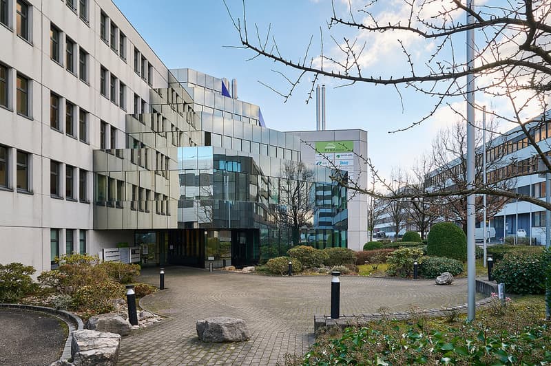 Multi-story office building with glass and concrete facade, surrounded by landscaped gardens and a paved walkway. There are trees and shrubs in the foreground, and a parking area is visible.