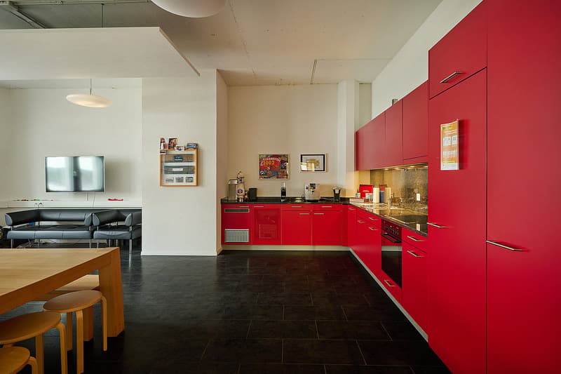 Modern kitchen with red cabinets, black countertops, stainless steel appliances including a dishwasher, and an open layout connecting to a living room area with a sofa and TV
