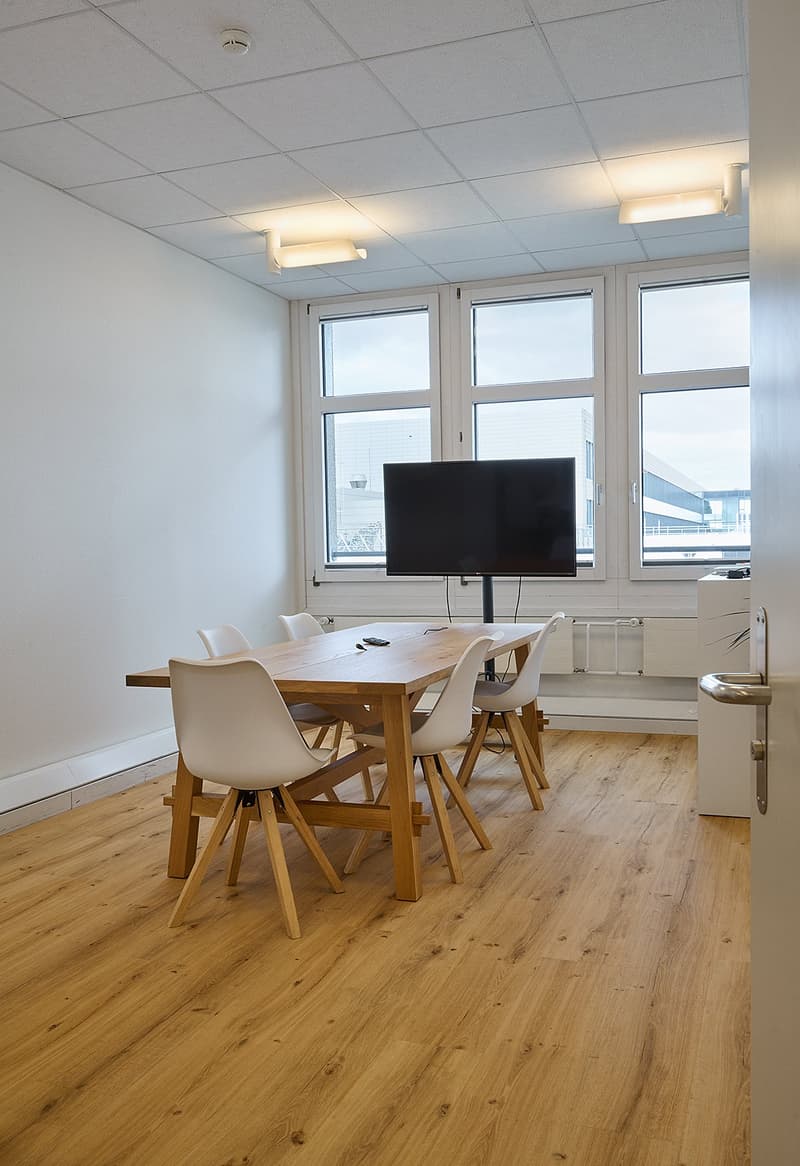 The image shows a modern office space with a large wooden table, white chairs, and a flat-screen TV mounted on the wall. The room has large windows that provide natural light, and the ceiling is equipped with recessed lighting. The floor is made of hardwo