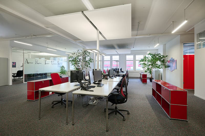 Large open office space with white walls, high ceilings, and large windows. Desks with computers and office chairs, red storage cabinets, and potted plants throughout the room.
