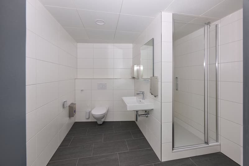Modern bathroom with white tiled walls, dark gray tile flooring, a toilet, a sink with a mirror, and a glass shower enclosure.
