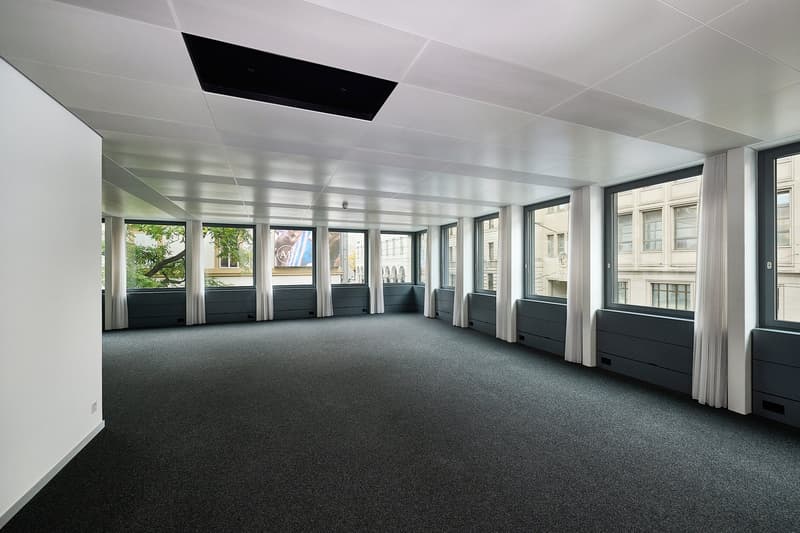 Large open office space with floor-to-ceiling windows, white walls, and dark gray carpeting. The room has a high ceiling with recessed lighting and appears to be designed for commercial or office use.
