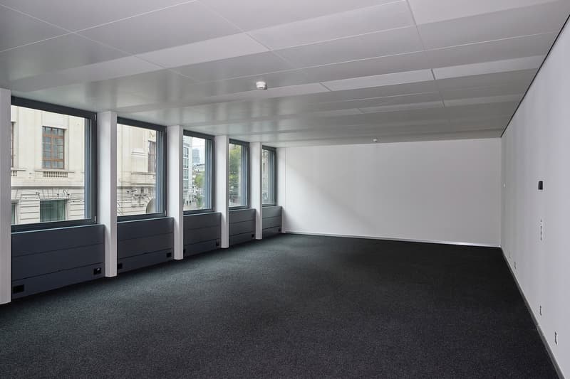 The image shows a large, open office space with a high ceiling and multiple windows along the walls. The room has a clean, modern design with white walls and a gray carpeted floor. There are several black storage units or cabinets lining the walls, and th