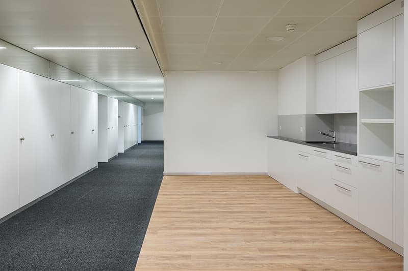 Large open office space with white walls, gray carpeted floor, and a kitchenette area with white cabinets and a stainless steel sink. The space has a modern and minimalist design.