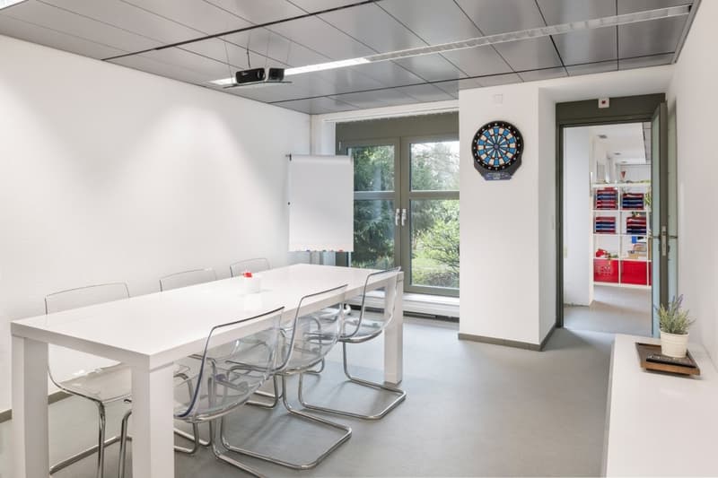 Long white table with transparent chairs, whiteboard on wall, projector on ceiling, dart board, door leading to another room