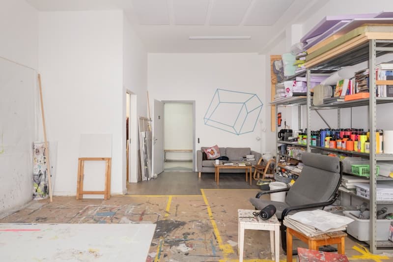 large room, white walls, wooden floor, shelves with various items, couch, table, wooden easels, paint splatters on the floor