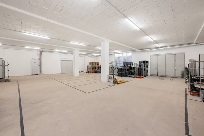 Empty warehouse with white walls, high ceiling, fluorescent lights, large doors, and storage equipment.