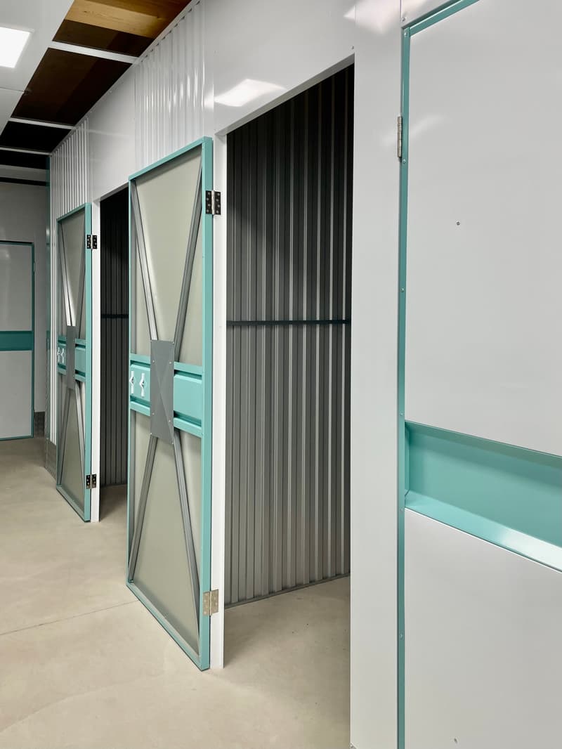 The image shows a storage unit with multiple compartments. The compartments have teal-colored frames and sliding doors made of a translucent material. The walls are white, and the floor is a light-colored concrete. The overall design appears to be modern