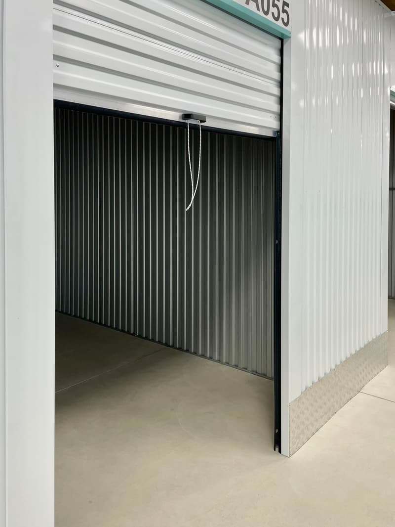 The image shows a storage unit with a white corrugated metal exterior and a black metal roll-up door. The unit appears to be located in a storage facility, with a concrete floor and a clean, organized interior visible through the open door.
