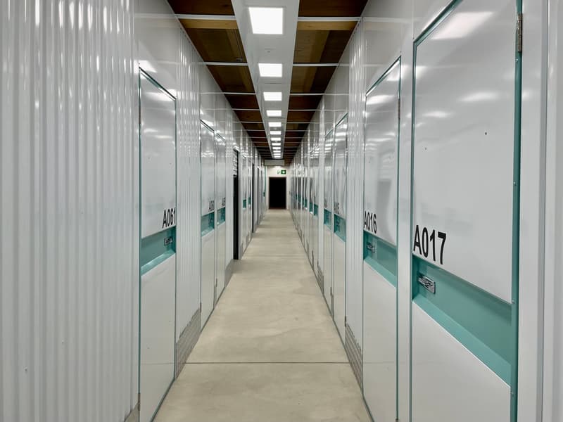 The image shows a long, narrow hallway with multiple storage units or lockers on both sides. The units are labeled with numbers A064, A016, and A017. The hallway has a concrete floor and the ceiling is lined with lighting fixtures. The overall appearance