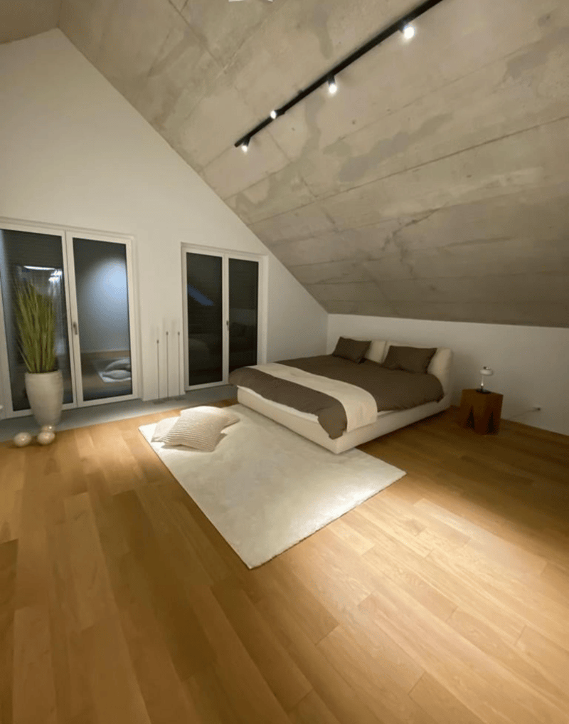 Master bedroom with two beds, white rug, wooden floor, white walls, glass sliding doors, mounted lamps, white plant vase