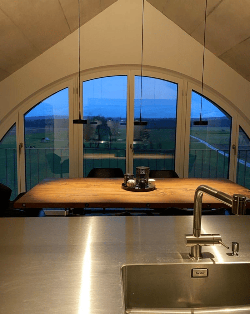 Dining area with long wooden table, 3 black chairs, metal balustrade, stainless steel sink, faucet, long windows with view to the landscape