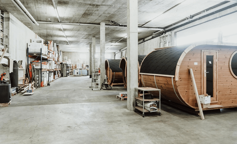 industrial-style warehouse, concrete floor, metal shelves, wooden pallets, trolley, storage boxes, two wooden barrels on stands, open windows