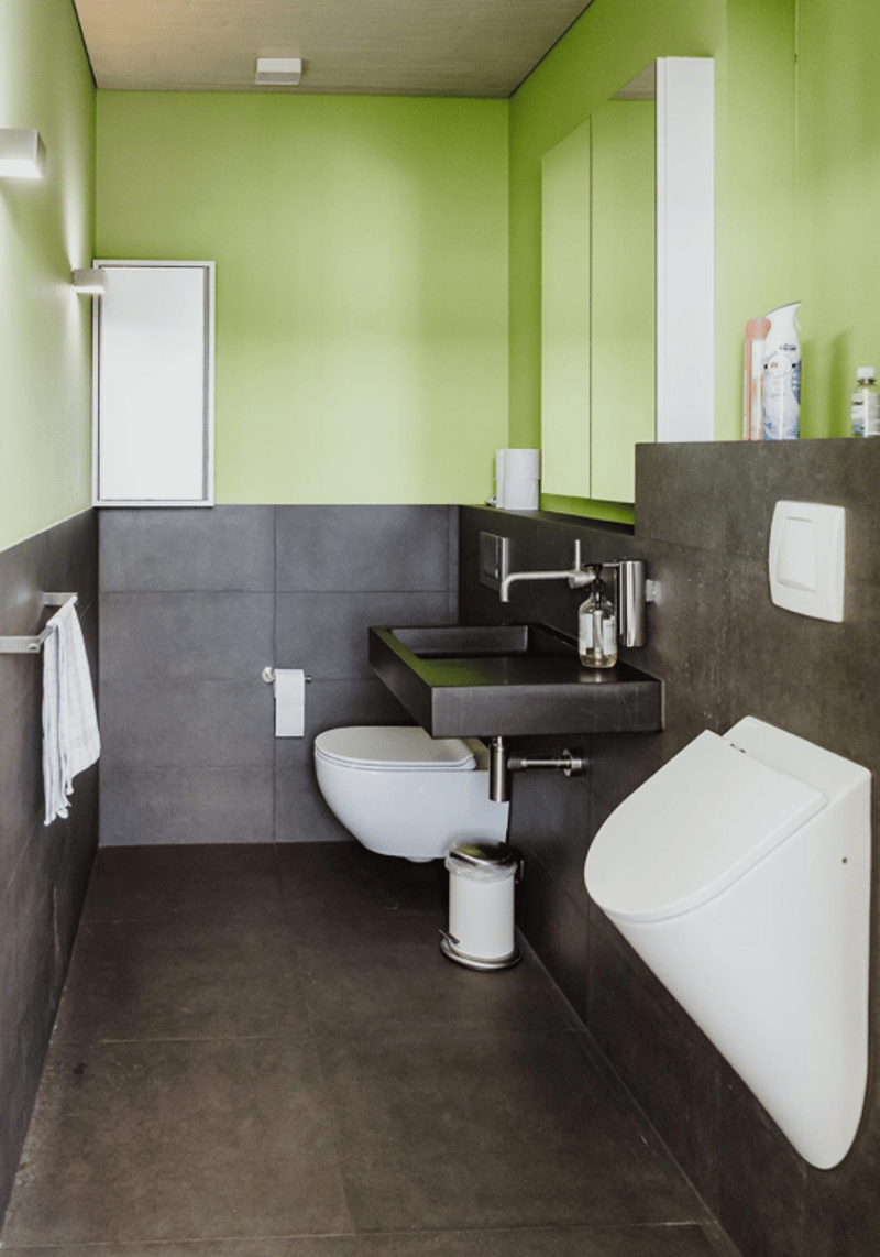 Compact bathroom with green walls, black sink, white fixtures, urinal, and a toilet with a trash bin