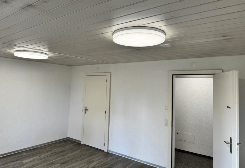 A room with white walls and wooden floor. The ceiling has two white ceiling lights. There is a white door and another open door. The right open door leads to a dark area.