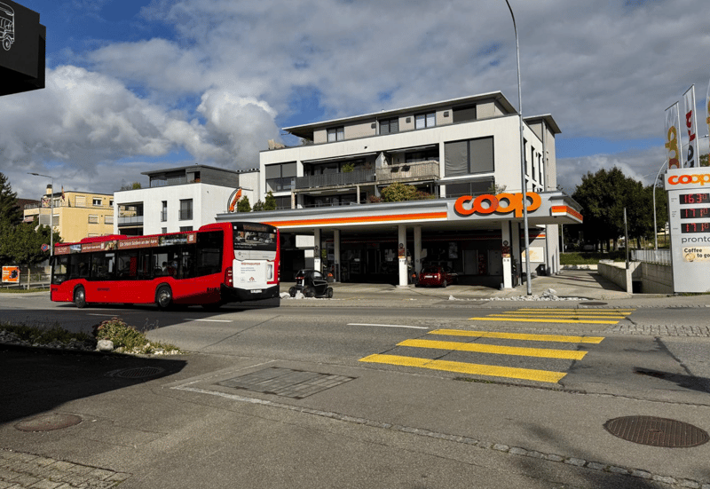 A gas station with the name 'COOP' is next to an apartment building. A red bus is driving by the gas station. A tree is on the left side, and some signs are near the road.