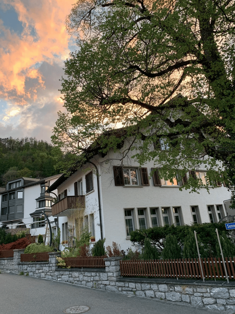 white facade house with brown wooden shutters, balcony on first floor, red roof, mountain in background