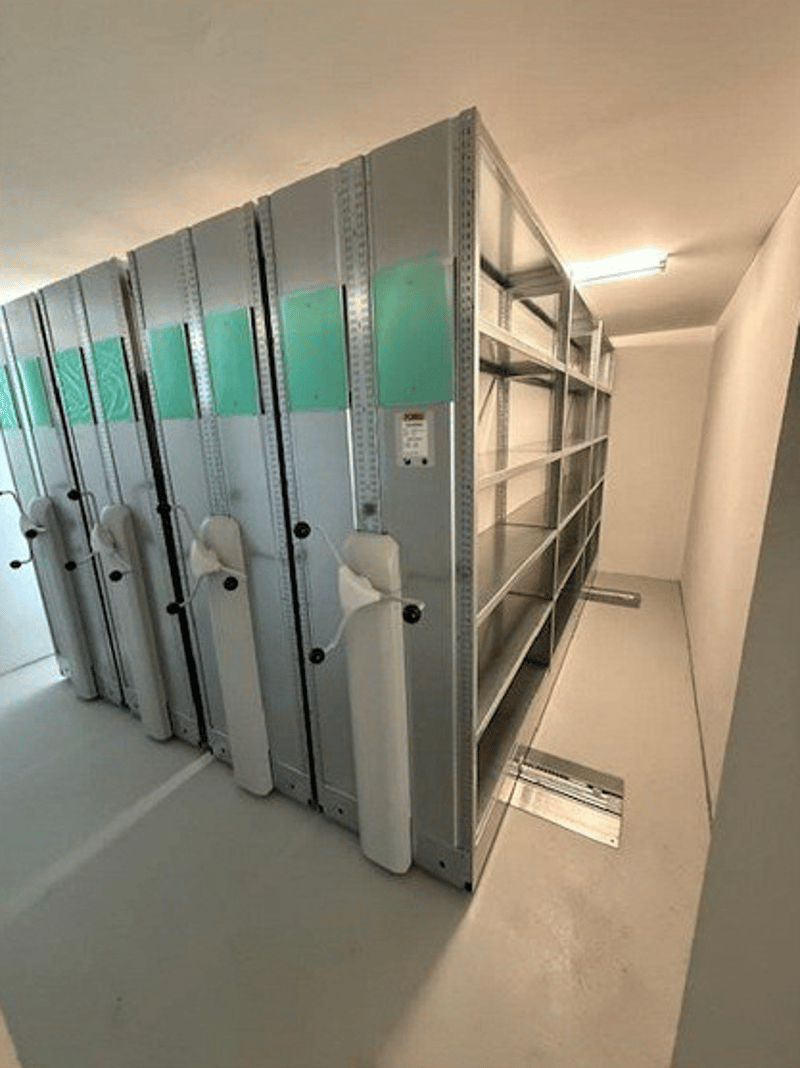 Storage facility with grey shelving, ceiling mounted lamps, white walls, smooth floor.