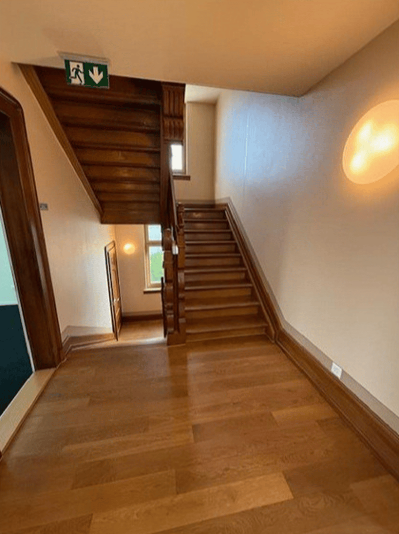 An interior hallway of a building with wooden staircases on both sides, white walls, and wooden flooring.