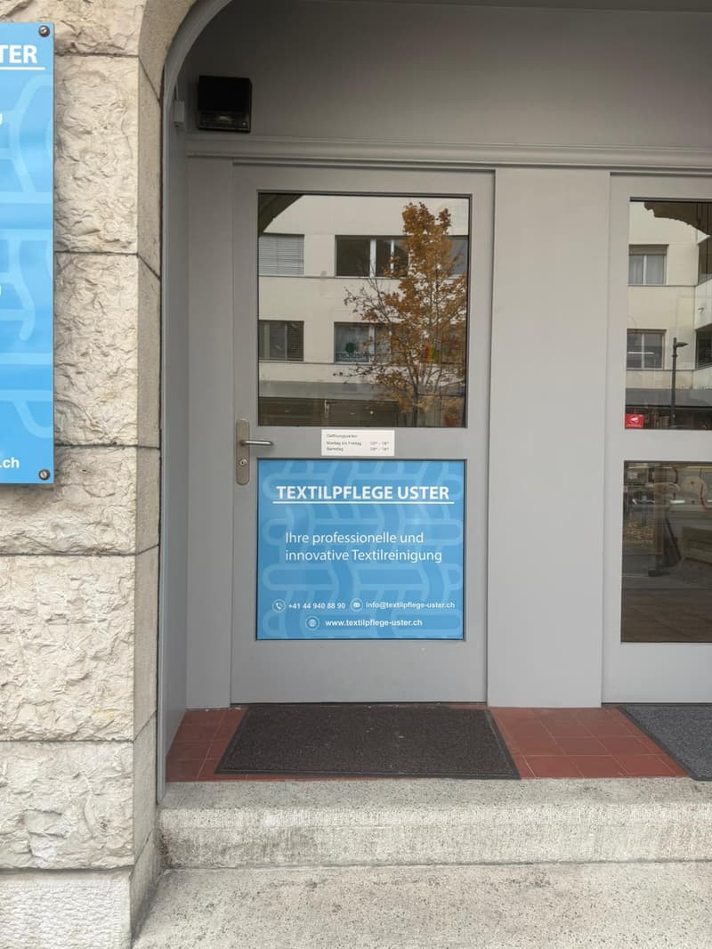 Building entrance, grey door with a sign and glass panel, text in German and English advertising professional and innovative textile cleaning services.
