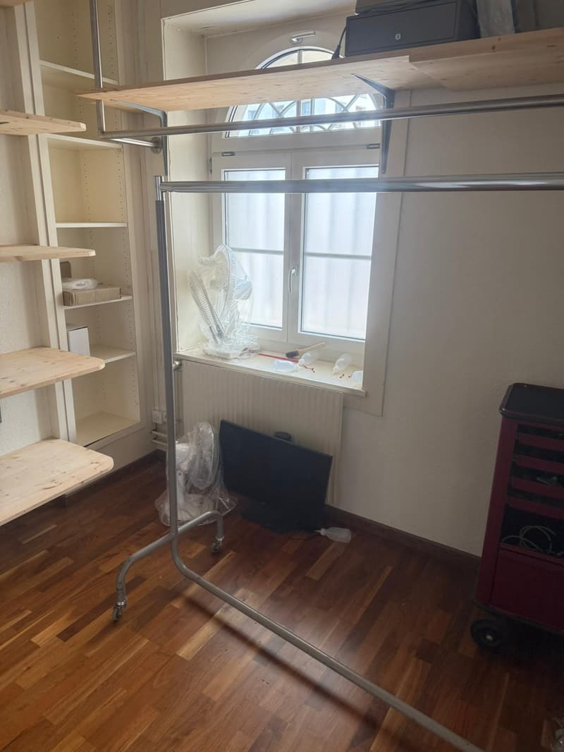 Room with white painted wall, a window with white painted frames, wooden floor, a metal cloth stand, a cabinet on the left, and a desk on the right