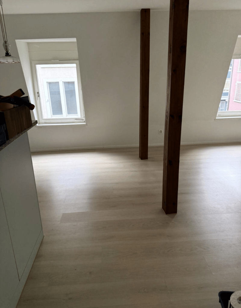 An empty room with wooden flooring, white walls, wooden columns, two windows, a countertop, and a cabinet. A lamp is hanging from the ceiling.