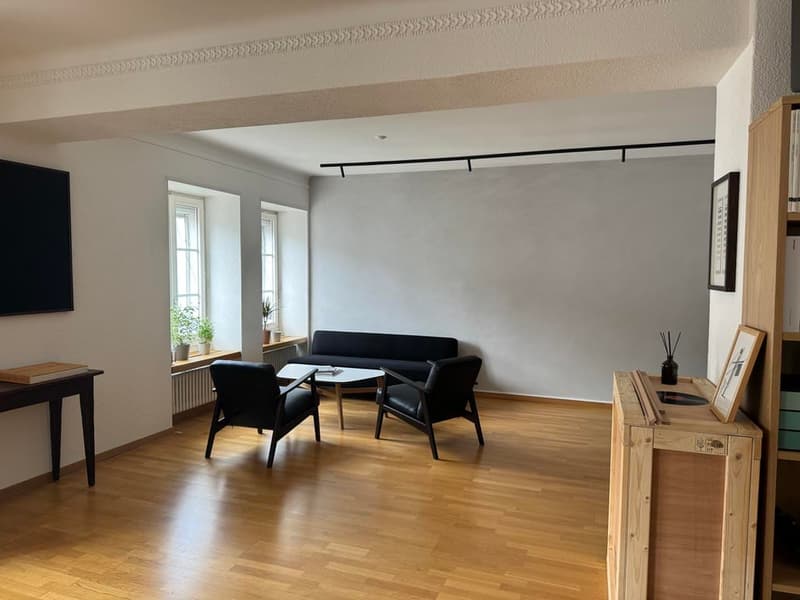 wood floor, two couches, round coffee table, two black chairs, shelf with books, two windows, a wall mounted tv, picture frame on wall