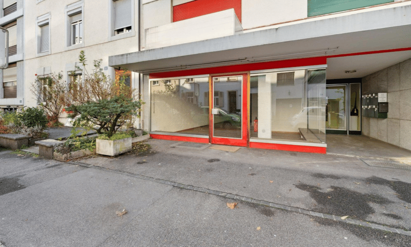 The entrance of an apartment building has large glass doors and windows. There are plants and greenery at the entrance and bicycles are parked on the side. A green car is parked in front.