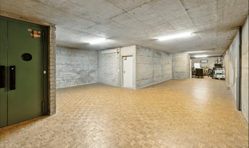 Empty basement room, concrete walls, wooden flooring, green door, white door, white ceiling with lighting, brick wall, no windows