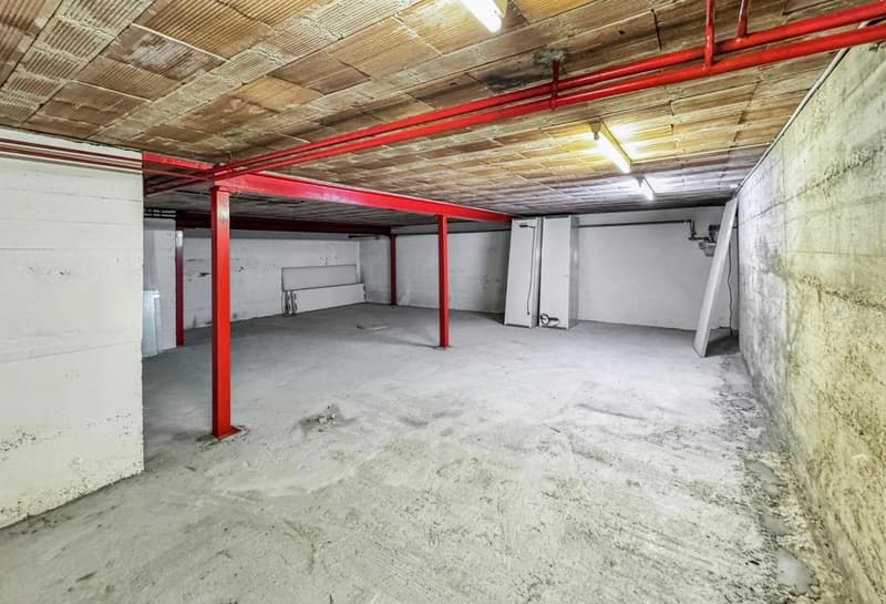 Concrete floor, red pillars, two electrical boxes, white walls, ceiling with lighting.