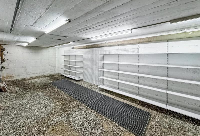 Empty storage room with two white shelves, metal grid on floor, white concrete walls, four ceiling lights.
