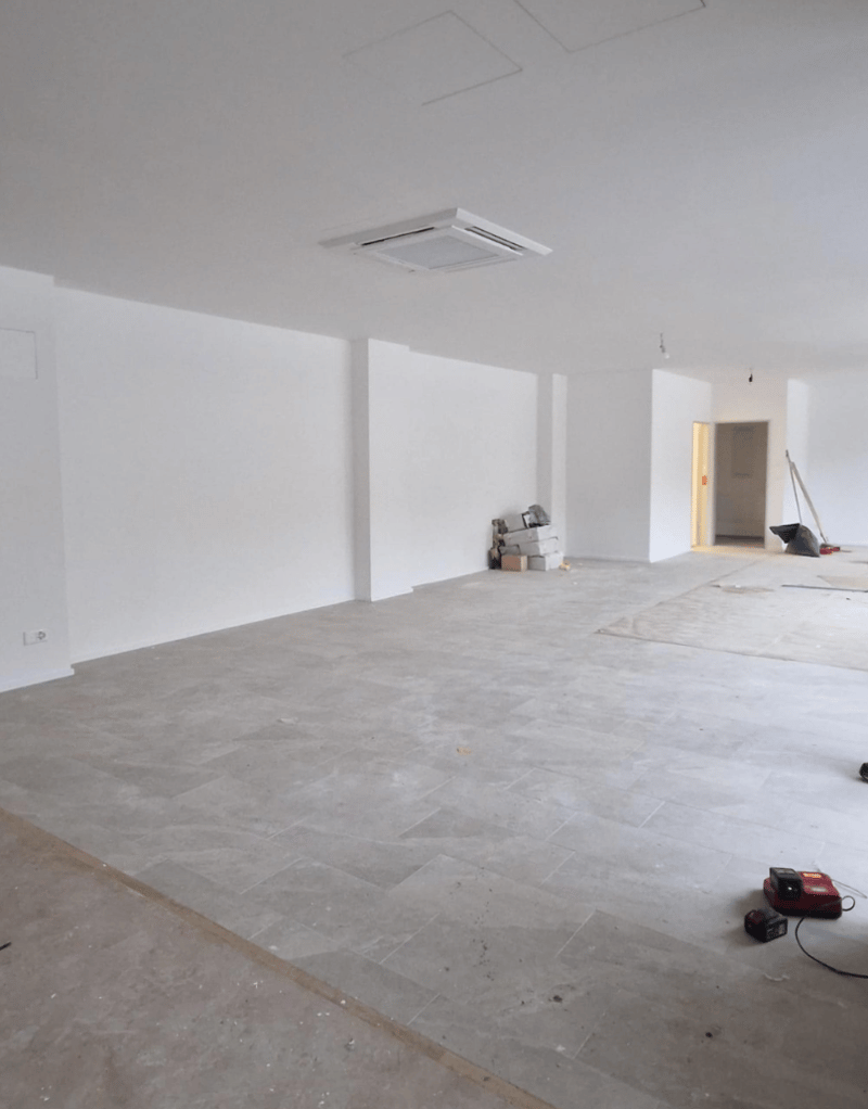 An empty room with white walls, grey tiled floor, air conditioner mounted on the ceiling, small items on the floor, and an electrical outlet.