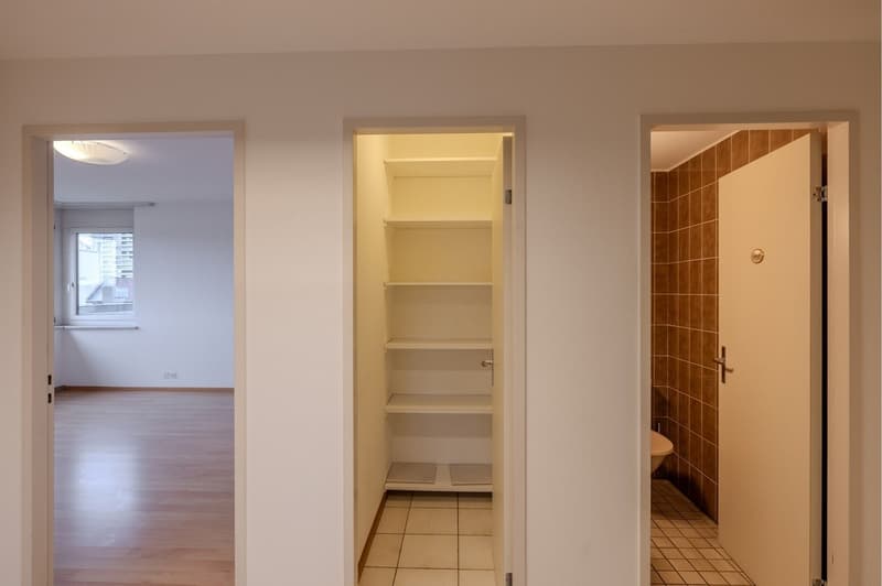 A hallway leading to a room with wooden floors and a window, an open door leading to an empty storage space, and an open door leading to a bathroom with tiled flooring.