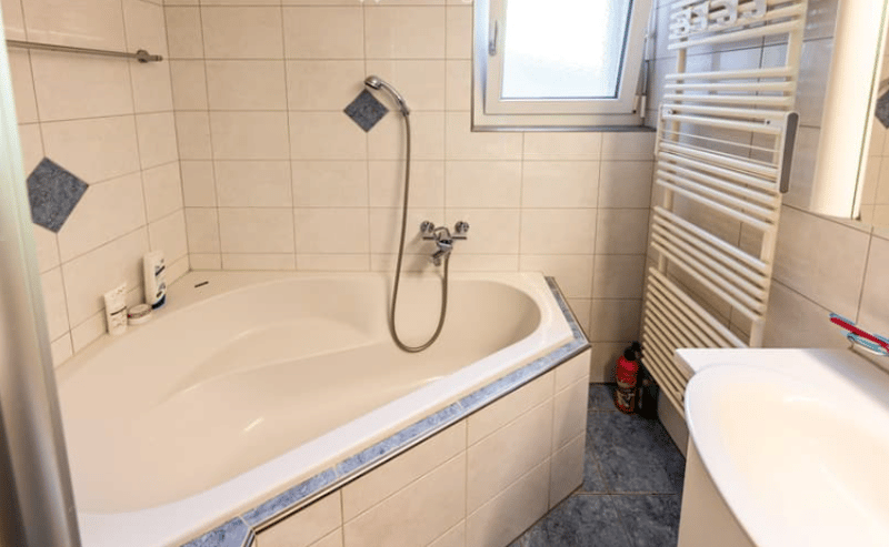 A bathtub is placed beside the sink. The bathroom has white tiled walls and floors. A towel rack and a toilet paper holder are mounted on the wall. A window provides natural light.