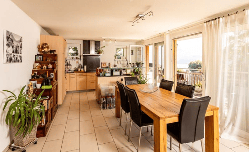 tiled floor, wooden table, black chairs, large windows, balcony, kitchen with black appliances, shelves with decor