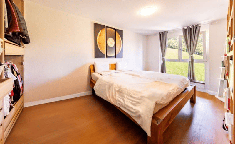 Bedroom with wooden bed, white linens, nightstands, white walls, and a large window with a grassy view outside.