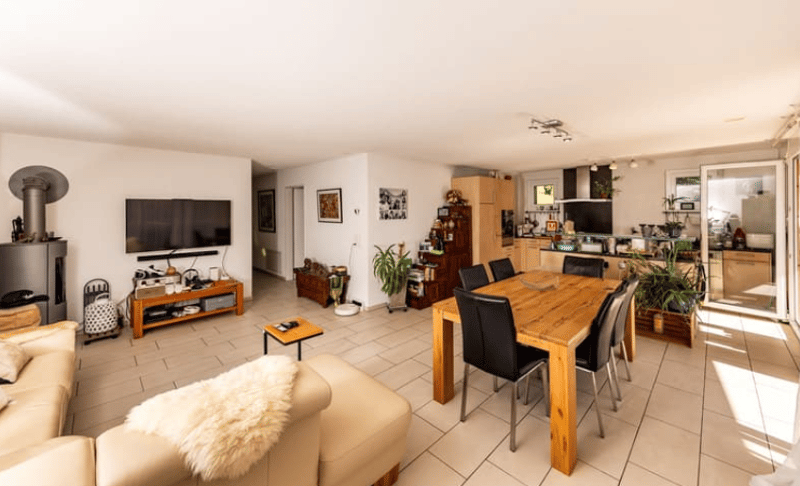 Living room with beige sofa, wooden dining table, black chairs, mounted television, shelves, fireplace, tiled floor.