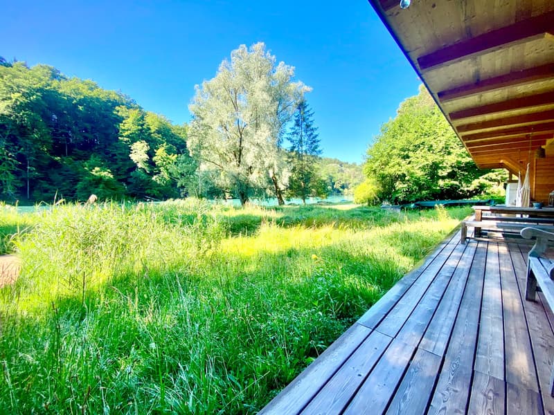 View of a large green grassy area with benches, trees, and a body of water, and a wooden deck with ceiling beams.