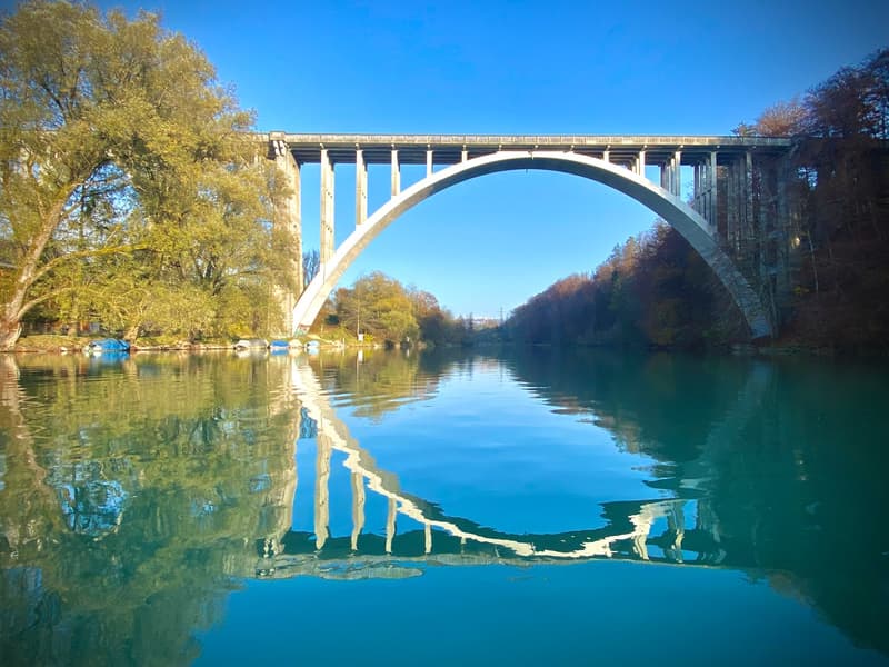 arch bridge over the river, reflecting on water, clear blue sky, trees in background