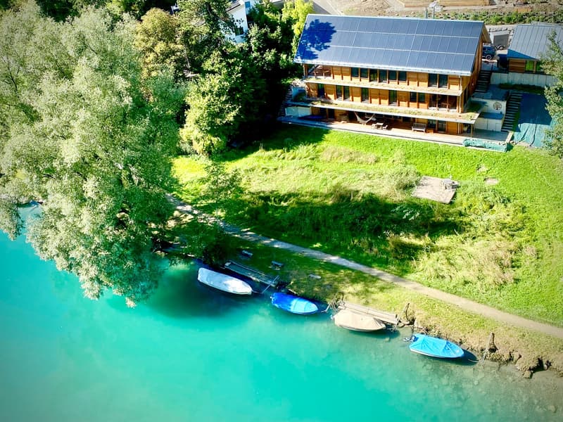Large house on lakefront, with solar panels on the roof, multiple boats moored on the lake, surrounded by lush green trees and grass.