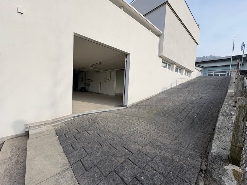 The garage entrance is made of white walls, a brick floor, and is sloped. The garage itself is empty and has an open door.