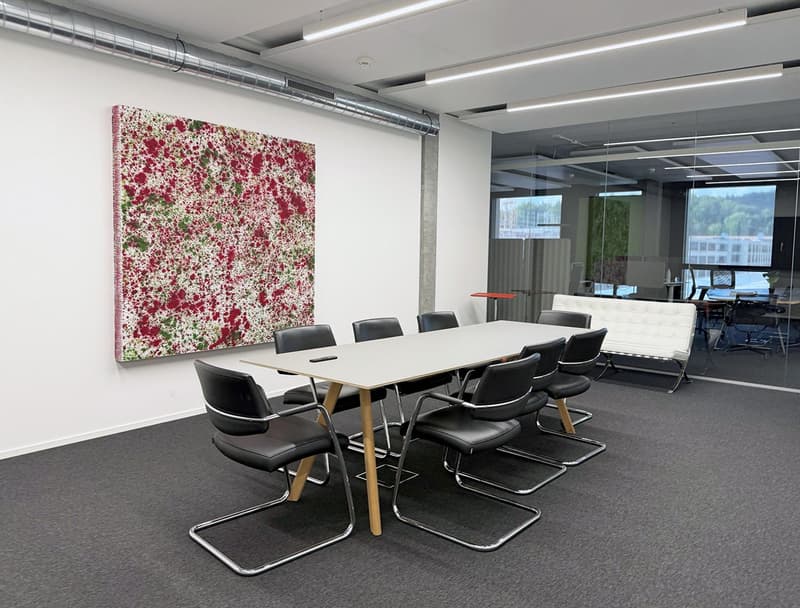 Modern office with a conference room setup. The conference room has a large white table surrounded by black chairs, a large colorful painting on the wall, and bright lighting from overhead lamps.