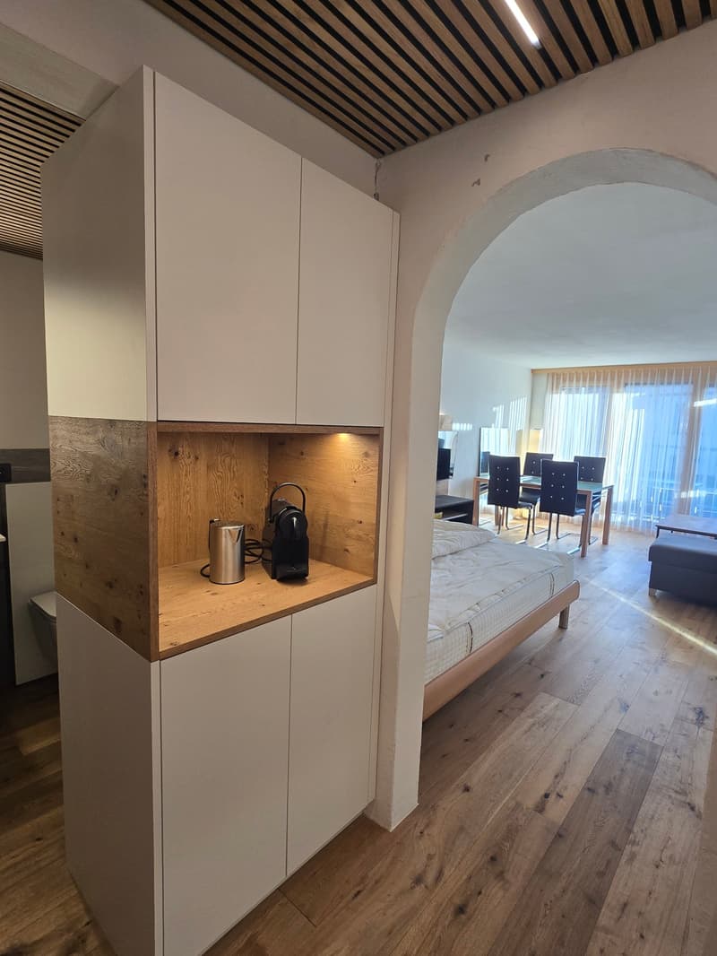 modern room with wooden floor, white cabinets, coffee machine, kettle, bed, dining table, chairs, ottoman, windows with curtains