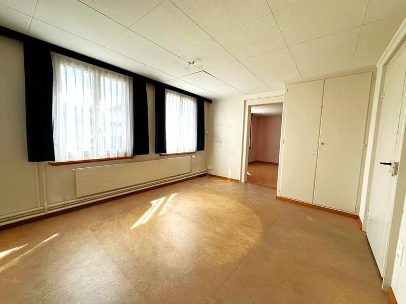 Empty room with wooden floor, white walls, large windows with black curtains, white radiators, and a white door.
