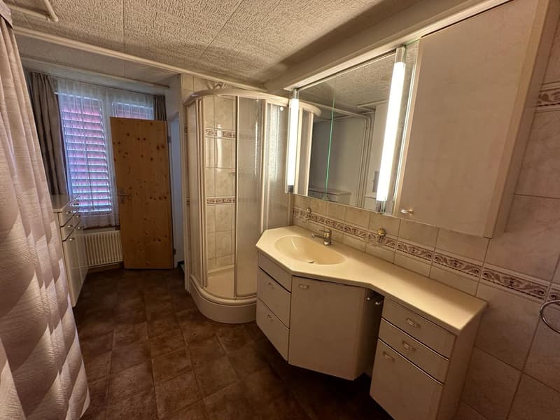 Bathroom with shower, sink, wooden door, radiator, tiled floor, wooden cabinet, mirror, and wooden door.