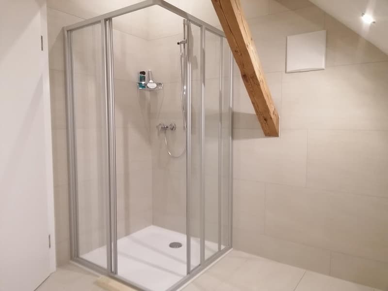 a shower with glass doors, tile floor, white wall, small square light, wooden beam, faucet, shower head, and a white power outlet